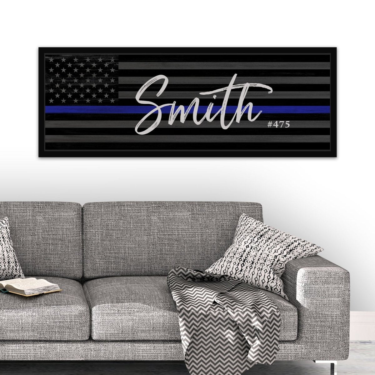 personalized thin blue line American flag wall art above gray sofa, custom police officer name sign “Smith #475”, black and white flag canvas with blue stripe, law enforcement appreciation home decor, modern living room display with framed police gift
