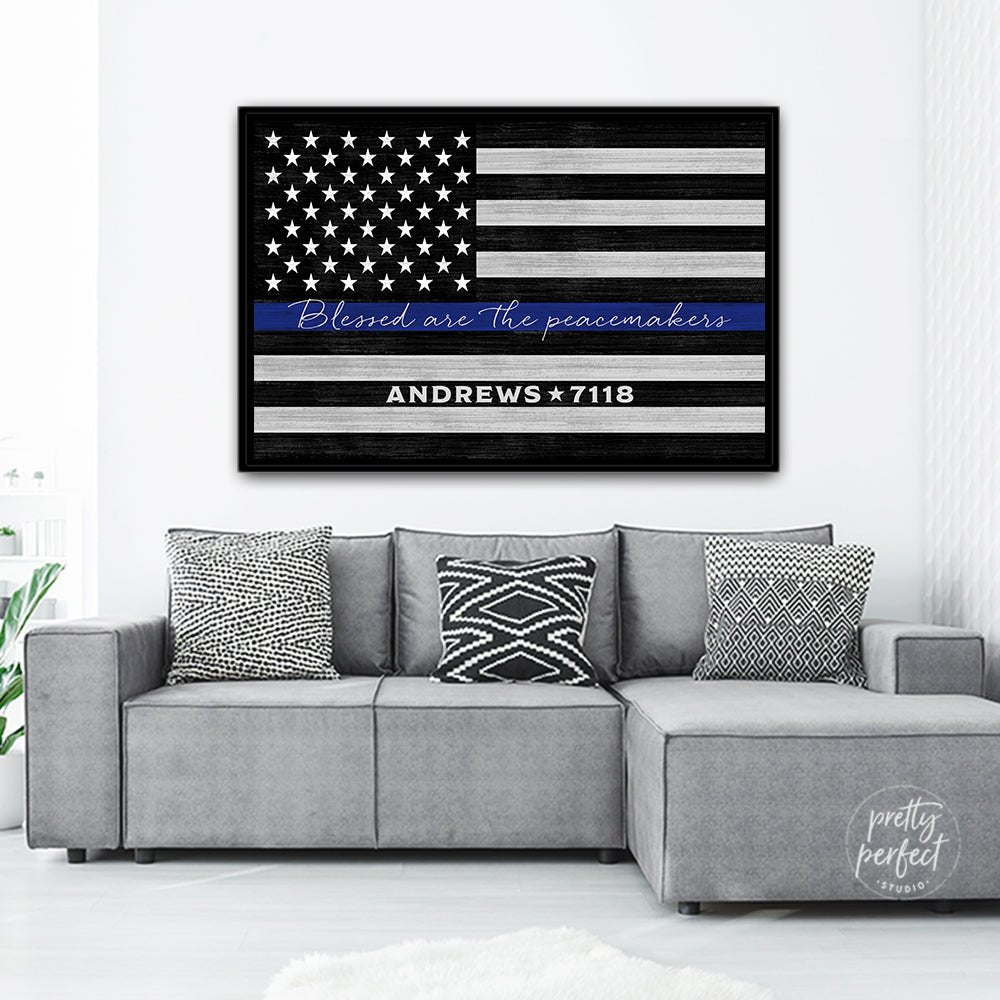 Personalized police officer wall art with thin blue line American flag design, black frame, distressed background, featuring “Blessed are the peacemakers” Scripture in white script across blue stripe and custom name and badge number displayed above modern gray sectional sofa