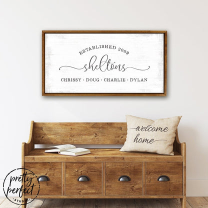 custom family name canvas wall art with last name, established date, and family members names in modern rustic style on lightly distressed background with teak frame, above entryway bench
