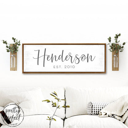 Personalized last name sign Henderson est 2010 farmhouse style custom family name wall decor above couch with rustic wood frame and greenery accents